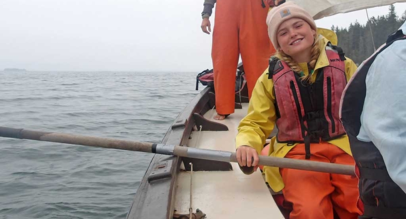 A person wearing a life jacket smiles as they hold the oar of a sailboat. 